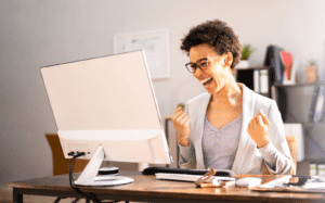 A delighted woman with curly hair and glasses, sitting at a desk and celebrating a moment of success in front of her computer, with a joyful fist pump.